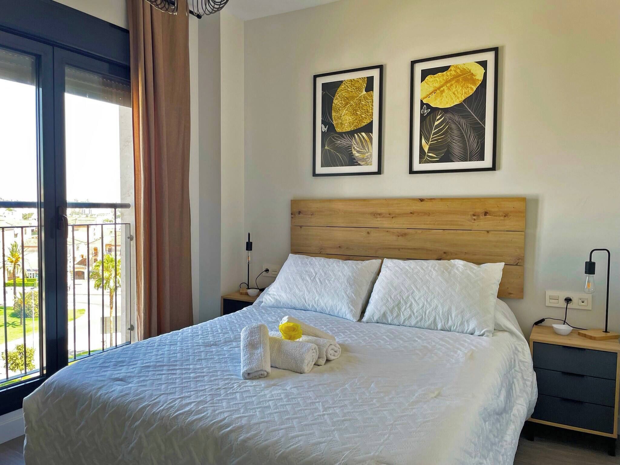 Ein Hotelzimmer mit einem großen Bett, einem hölzernen Kopfteil, zwei gerahmten Kunstwerken an der Wand, einem Nachttisch mit einer Lampe und Blick ins Freie durch ein großes Fenster.
