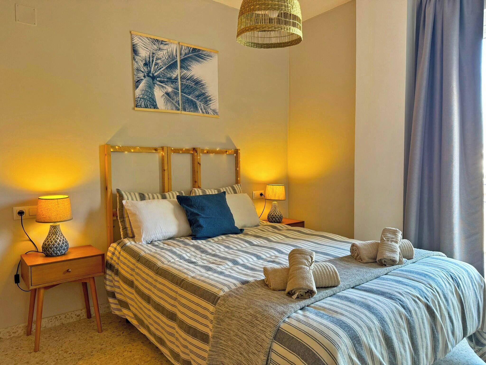 Een slaapkamer met een gestreepte spreideken, twee bedlampen, een hanglamp en een wanddecoratie van een palmboom.