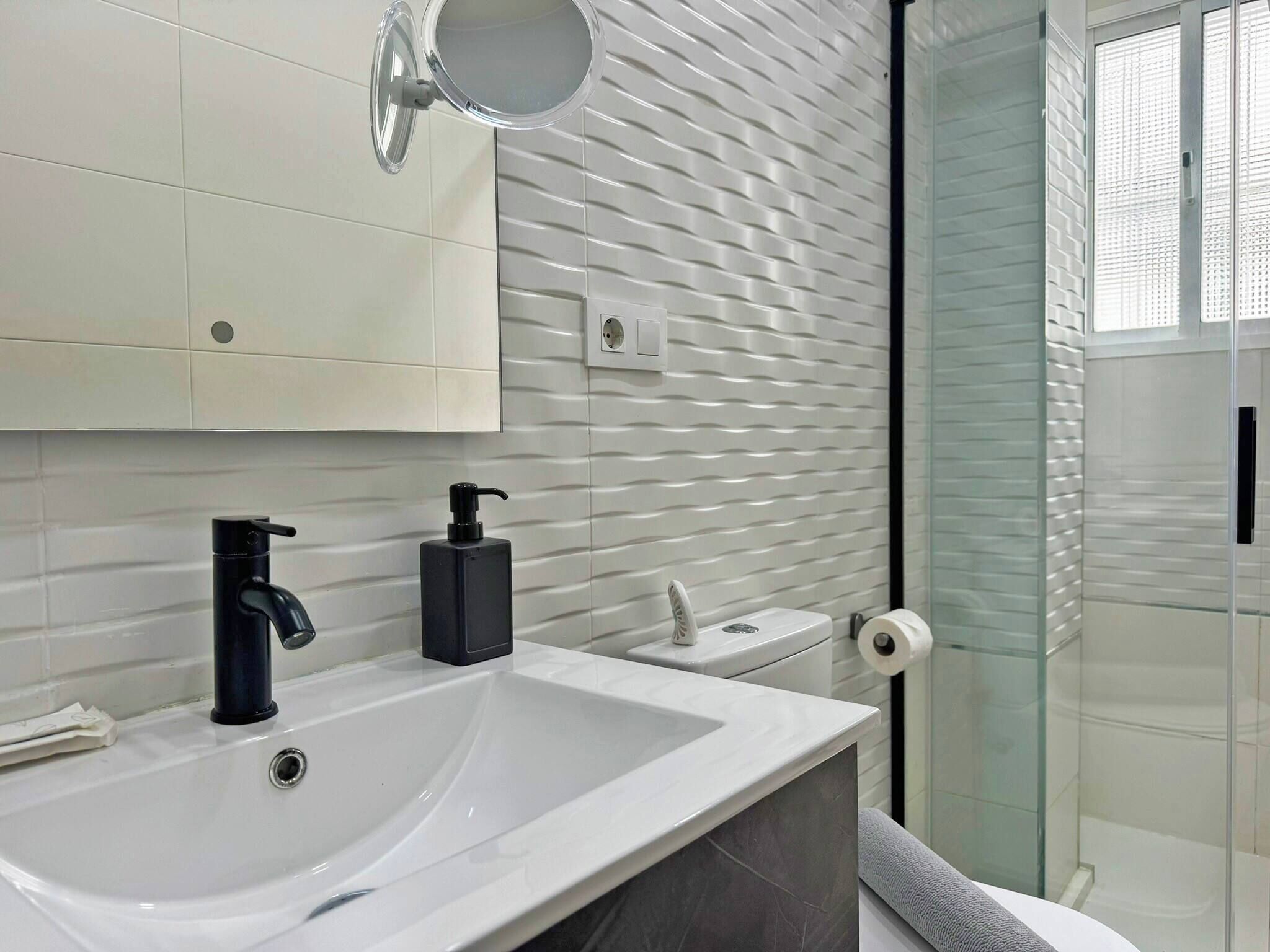 A modern bathroom with a white sink, black faucet, and soap dispenser. There is a toilet with a paper towel holder and a glass shower enclosure.