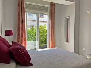 Ein Schlafzimmer mit Bett, Nachttisch, Lampe und Blick auf einen Balkon mit Grünpflanzen.
