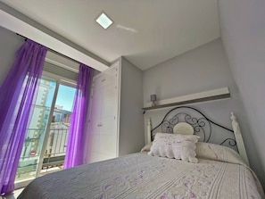 Un dormitorio con cama, ventana con cortinas moradas, luz de techo y vista de edificios.