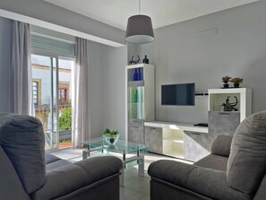 A modern living room with a glass coffee table, grey sofas, a TV mounted on the wall, and a balcony with curtains.