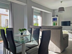 A modern living room with a glass dining table, grey sofa, and a TV mounted on the wall.