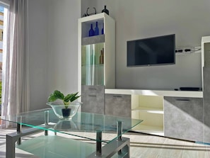 Ein modernes Wohnzimmer mit einem Glastisch, einem flachbildfernseher an der Wand und einer schlichten Kommode mit blauen Vasen.