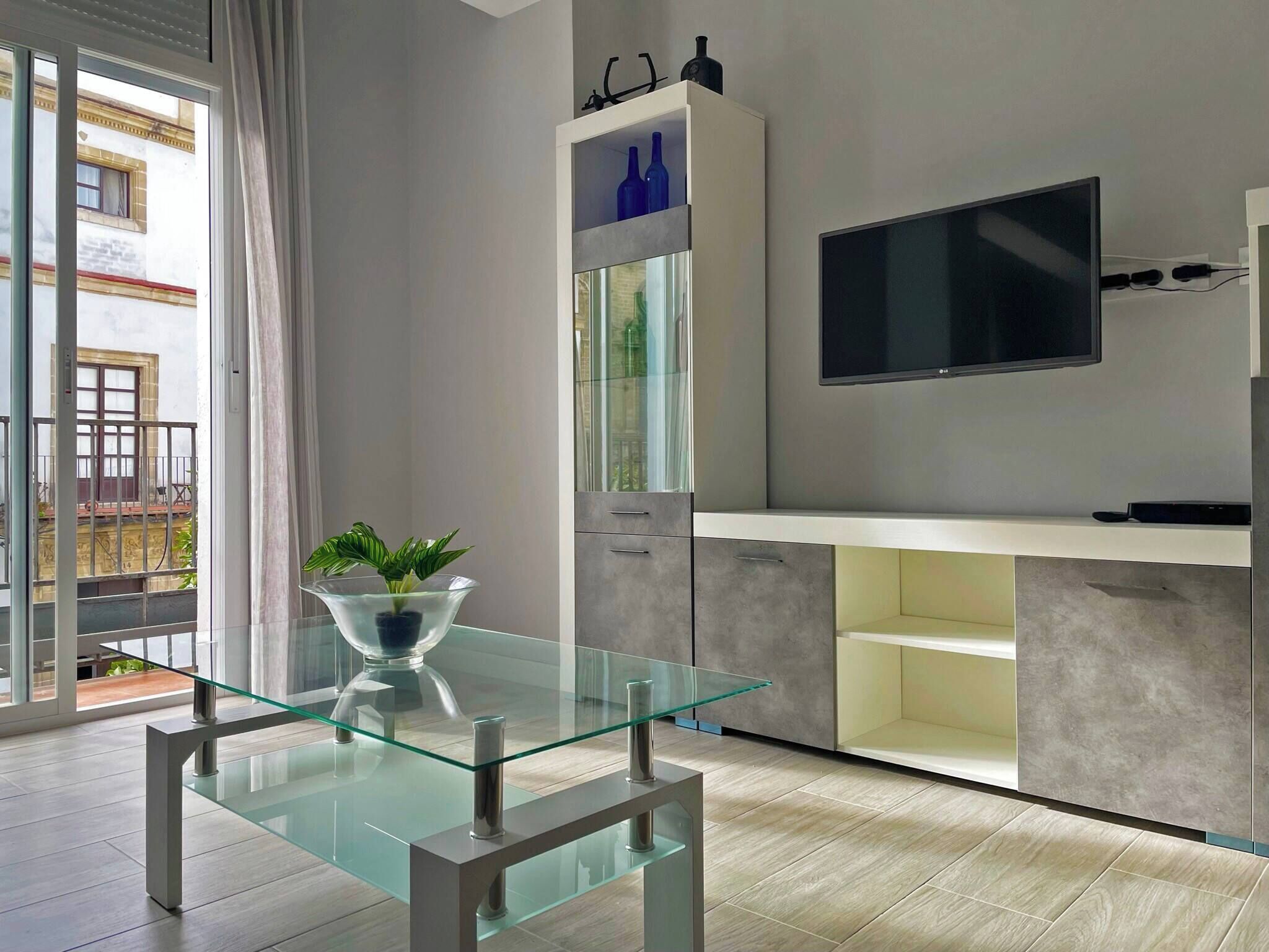 A modern kitchen with a glass-top table, a built-in TV, and a balcony with a view of the city.