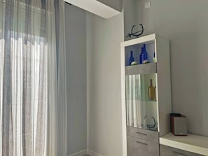 Una habitación moderna con un ventanal grande, un mueble blanco con puertas de vidrio y un altavoz pequeño de madera.