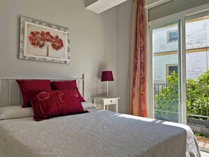 A bedroom with a bed, a nightstand, a lamp, a framed artwork, and a view of greenery outside. - Apartamento La Catedral - Ole Solutions (Jerez de la Frontera)