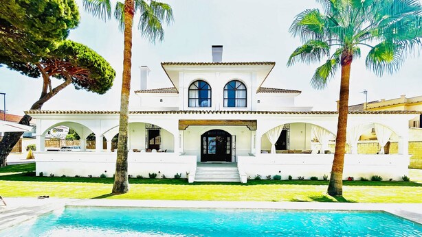 Une grande maison blanche avec une piscine devant, entourée de palmiers.