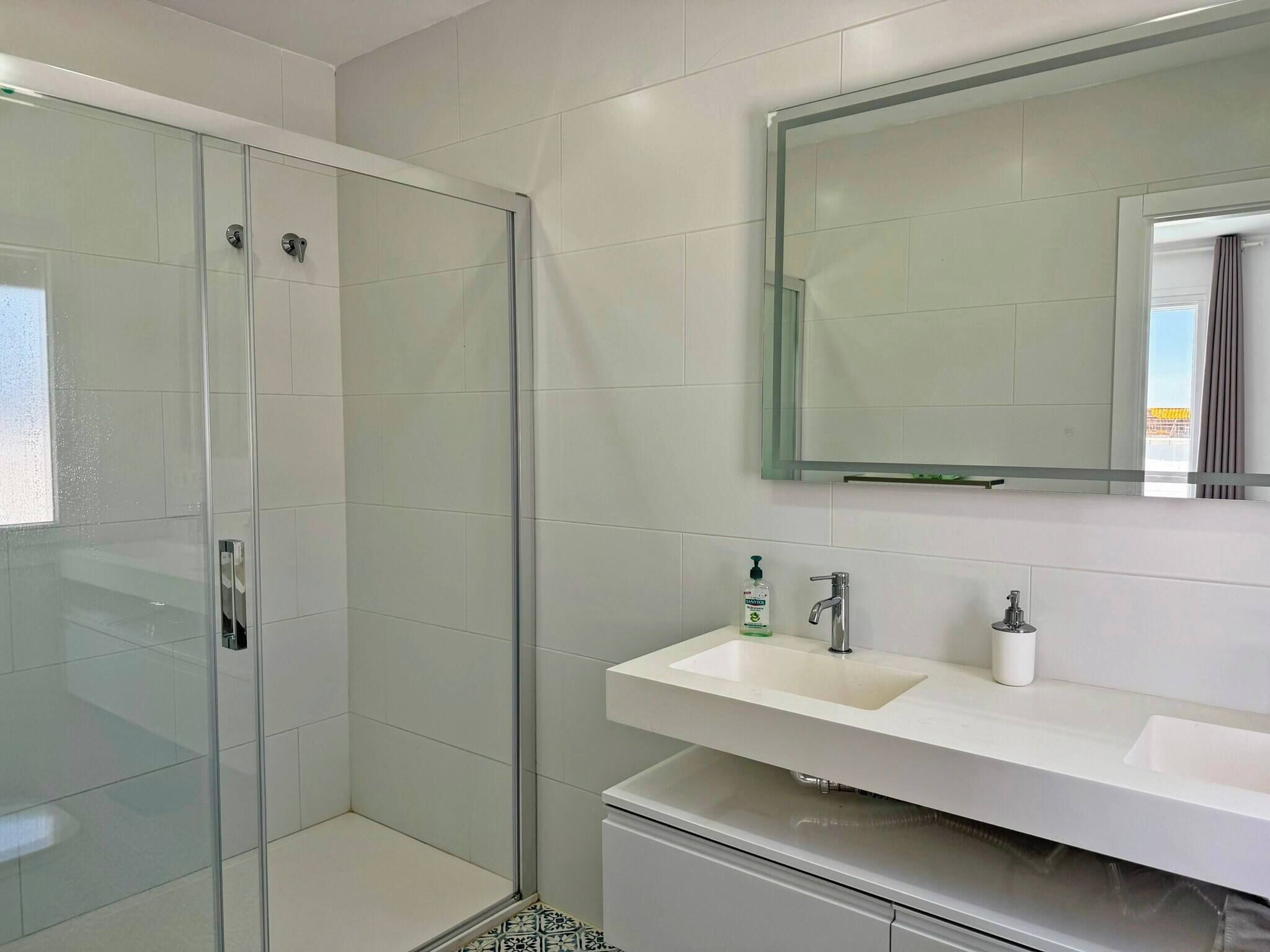 Een moderne badkamer met een glazen douchecabine, een witte wastafel met een spiegel erboven en een betegelde vloer.