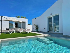 A modern outdoor pool area with a clear blue pool, a covered seating area with lounge chairs, and a white building with large windows.
