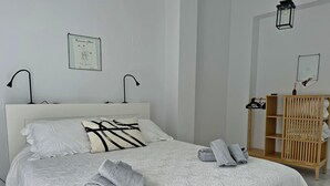 A neatly made bed with a white and patterned bedspread, two pillows, and rolled towels. There is a wooden wardrobe with a mirror and a framed picture on the wall. - Apartamento San Miguel - Ole Solutions (Jerez de la Frontera)