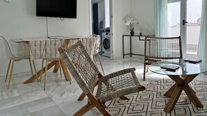 A modern living room with a glass coffee table, wicker chairs, a flat-screen TV, a washing machine, and a view of the outside through a glass door. - Apartamento San Miguel - Ole Solutions (Jerez de la Frontera)
