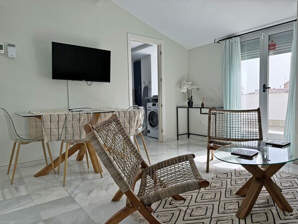 A modern living room with a glass coffee table, wicker chairs, a flat-screen TV, a washing machine, and a view of the outside through a glass door.
