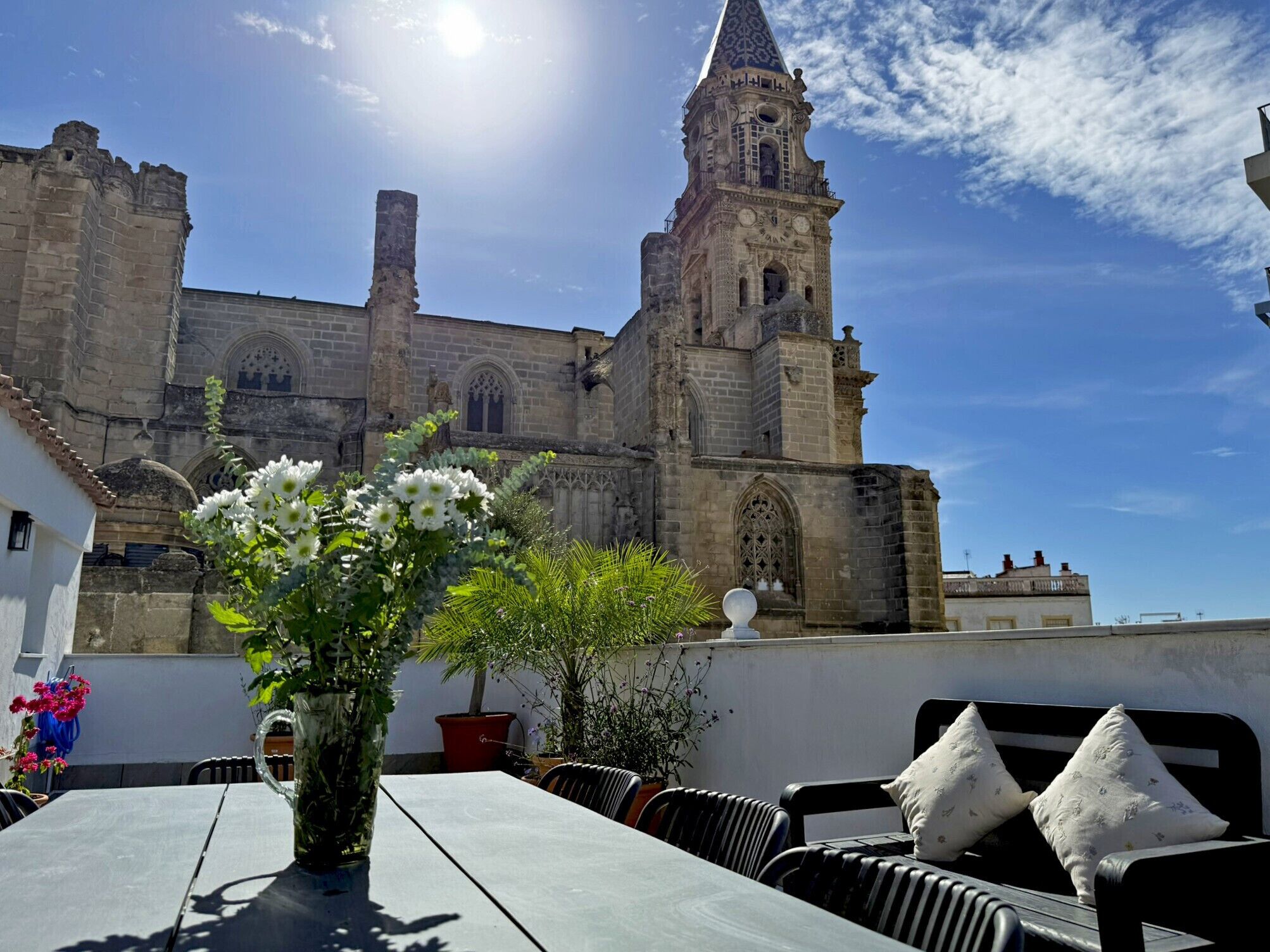 Un patio con una mesa y sillas, un jarrón con flores y un edificio histórico con una torre al fondo.