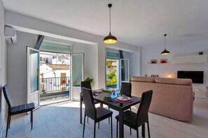 Ein modernes Wohnzimmer mit einer Couch, einem Esstisch mit Stühlen, Pendelleuchten und einem Fernseher an der Wand.