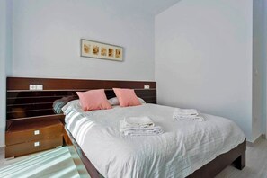 Ein ordentlich bezogenes Bett mit weißer Decke und rosafarbenen Kissen, ein hölzerner Kopfteil und ein kleiner Nachttisch mit Schublade.