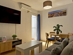 A living room with a flat-screen TV, a dining table with chairs, a sofa, a potted plant, and a painting on the wall.