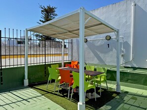 Un espace extérieur abrité avec des chaises vertes et orange, une table et un mur blanc orné d’un panneau indiquant un numéro.