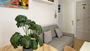 Una sala de estar con un sofá, una planta en maceta, un cuadro, un estante con adornos y una puerta con número.