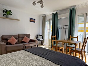 Une pièce comprenant un lit, un canapé, une table à manger avec des chaises, un luminaire suspendu, une plante en pot et une fenêtre avec des rideaux.