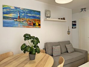 A living room with a painting, a potted plant, a sofa, a table, and a shelf with decorative items.