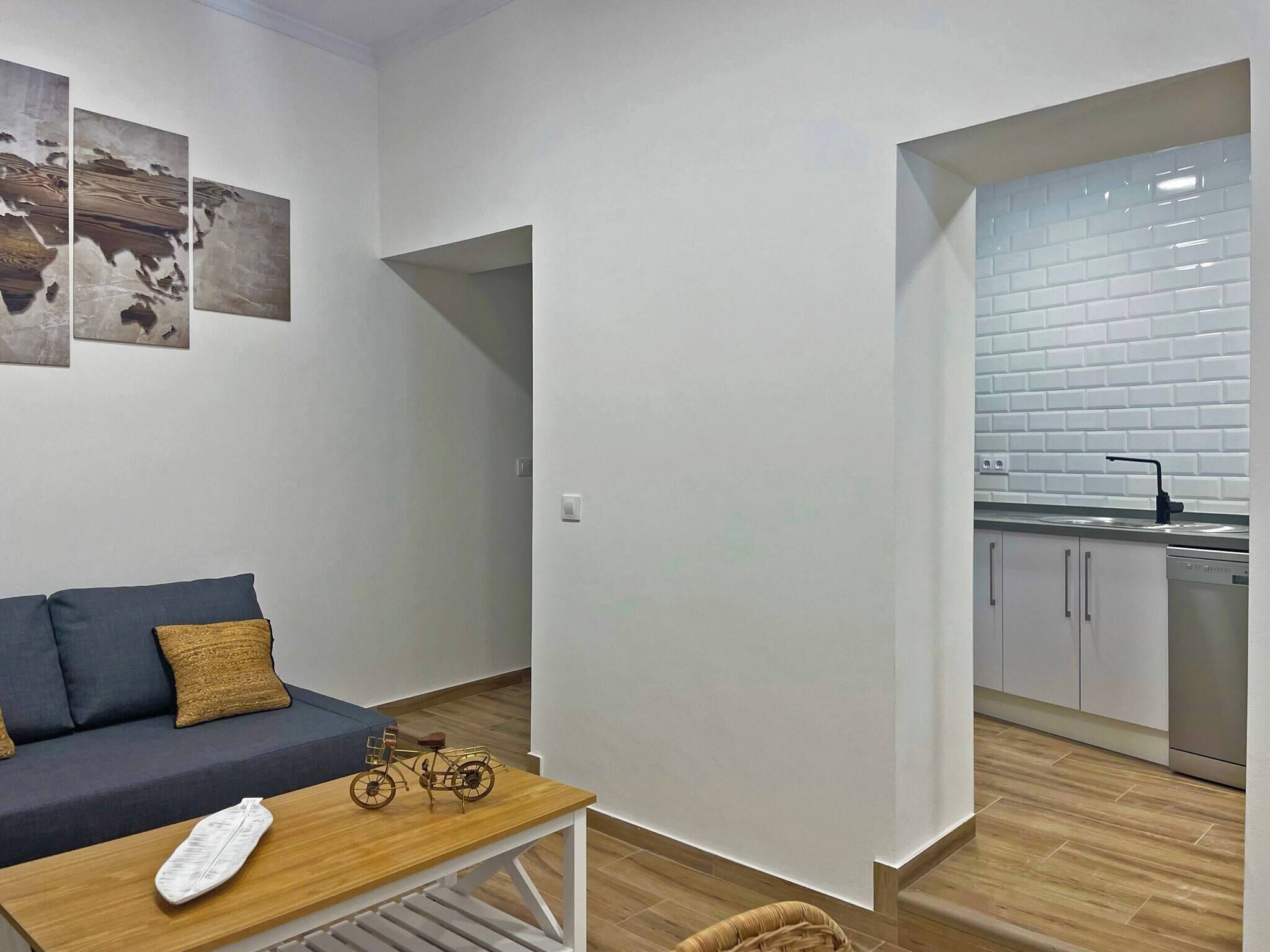 A modern apartment with a living area featuring a sofa, a wooden coffee table, and a small bicycle. A kitchen with white subway tiles and stainless steel appliances is visible through an open doorway.