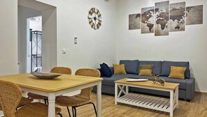 Una sala de estar con comedor, un sofá gris, sillas de mimbre, una mesa de centro de madera y un reloj fijado a la pared.