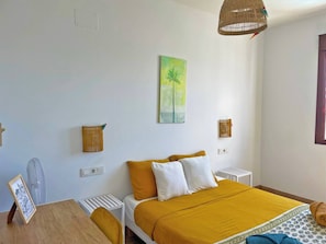 Una stanza con un letto, un tavolo, una sedia, un ventilatore e un quadro appeso al muro.