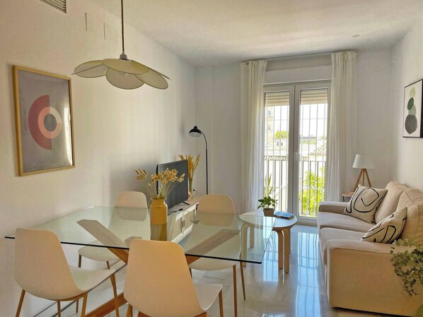Una sala de estar moderna con una mesa de comedor de vidrio, sofás blancos y un amplio ventanal con cortinas.