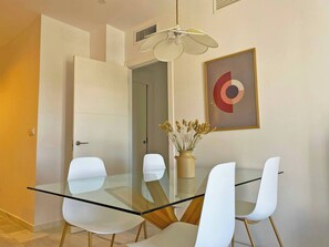 A modern dining area with a glass table and white chairs, a ceiling fan, and a framed abstract painting. - Apartamento El Carmen - Ole Solutions (Jerez de la Frontera)