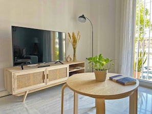 Een moderne woonkamer met een flatscreen televisie op een houten kast, een ronde houten tafel met een potplant en boeken, en een vloerlamp.