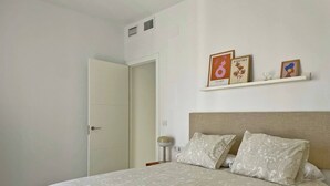 A bedroom with a bed, a bedside table, a nightstand, a wall-mounted shelf with framed pictures, and a door. - Apartamento El Carmen - Ole Solutions (Jerez de la Frontera)