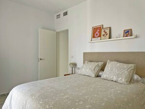 Un dormitori amb un llit, una taula de nit, una petita estanteria penjada a la paret amb fotos emmarcades i una porta.
