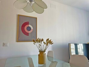 Una sala de reuniones moderna con una mesa de vidrio, un jarrón amarillo con plantas secas, una obra de arte abstracto enmarcada y un ventilador de techo.