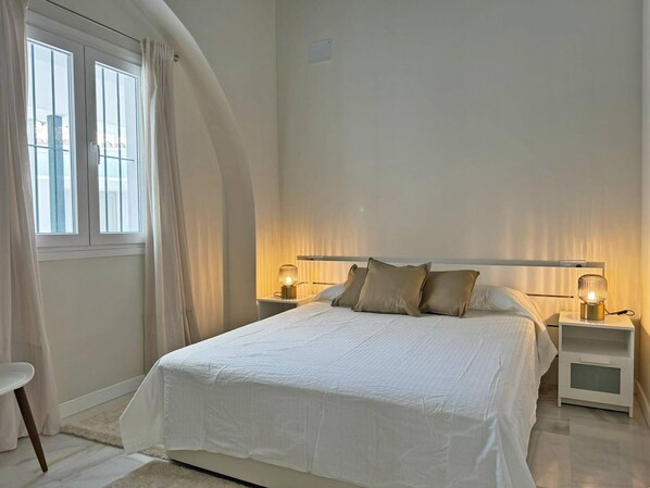 A bedroom with a large bed, two bedside tables with lamps, a window with curtains, and a view of the sea.