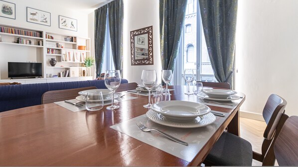 A dining area with a wooden table set for four, surrounded by chairs. There are bookshelves and a television in the background.