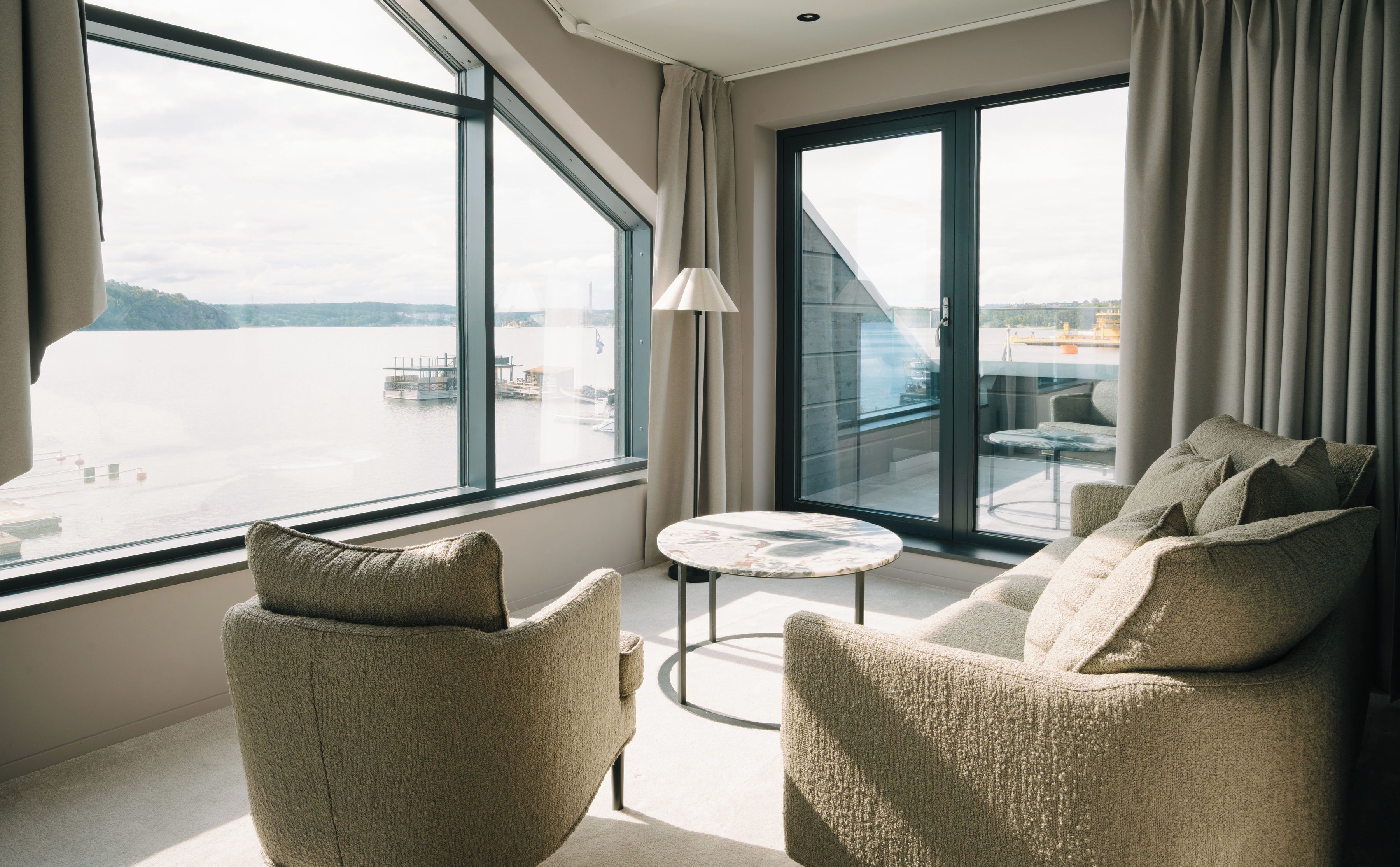 Ein modernes Hotelzimmer mit einem großen Fenster und Blick auf ein Gewässer sowie eine Anlegestelle. Ein runder Tisch und zwei Sessel mit Kissen sind vorhanden.