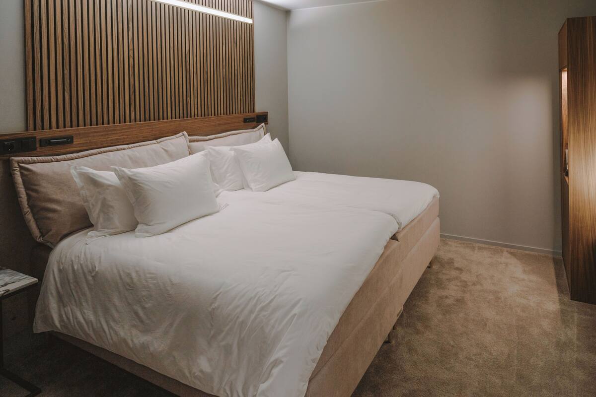 A neatly made bed with white linens in a room with a wooden headboard and a neutral wall.
