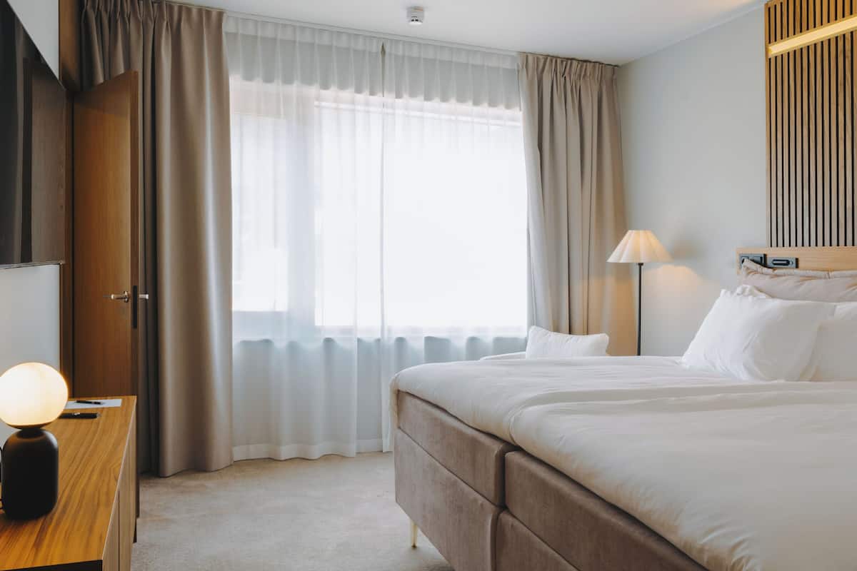 A hotel room with a large bed, a desk with a lamp, a window with curtains, and a bedside table.