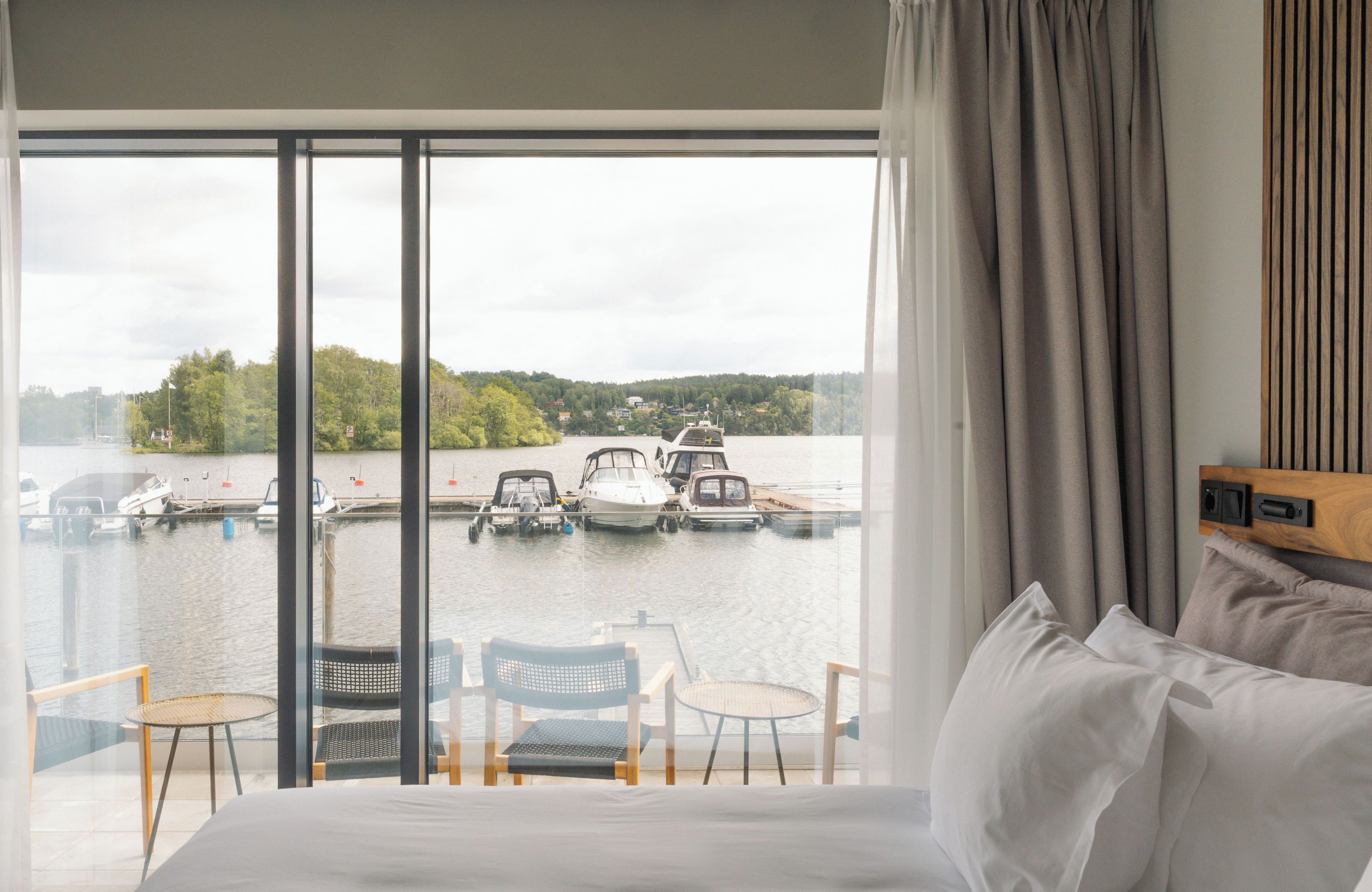 Habitación de hotel con un gran ventanal corredero que da a un balcón con vistas a un puerto deportivo con barcos. Hay dos sillas y una mesita en el balcón.