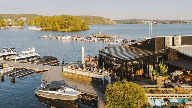 A marina with boats docked, a covered seating area, and a view of the water and distant land. - Season Hotel (Ekerö)