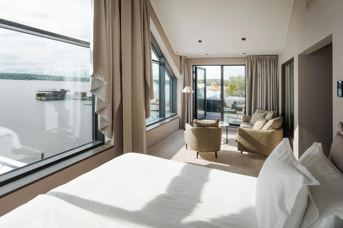 A hotel room with a large window, a bed, two armchairs, and a small table.