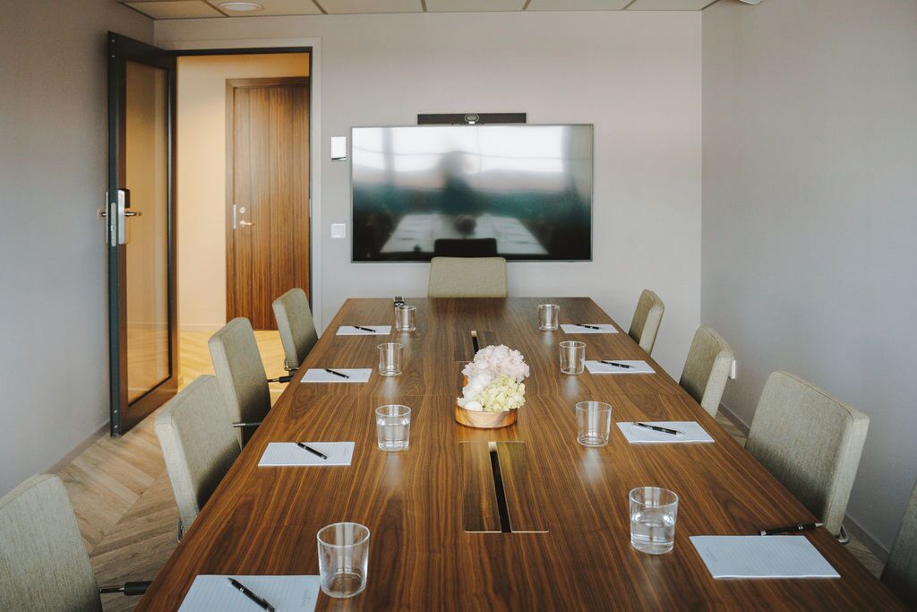 A conference room with a long wooden table, chairs, a TV screen, and a floral centerpiece.