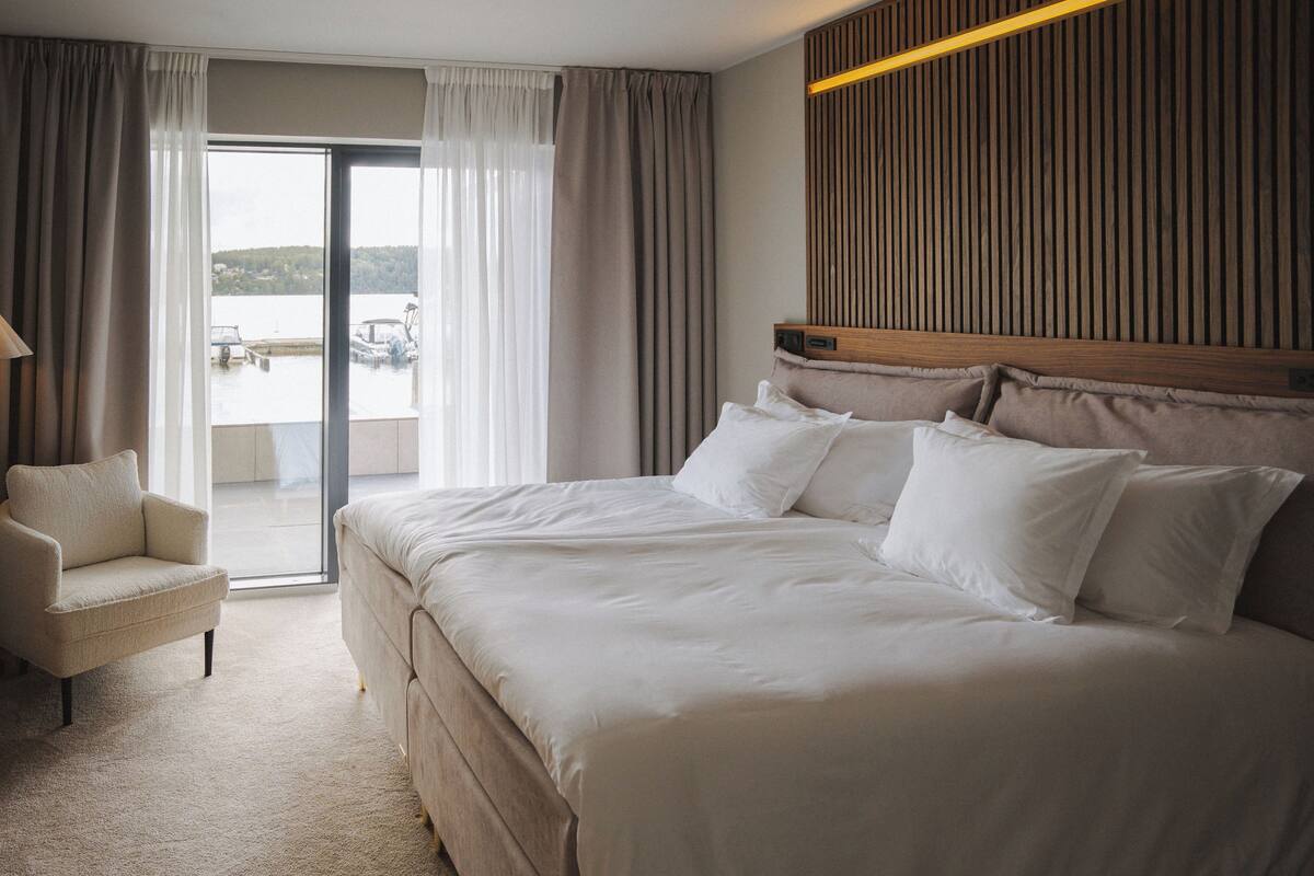 A hotel room with a large bed, a chair, and a view of a body of water through a sliding glass door.