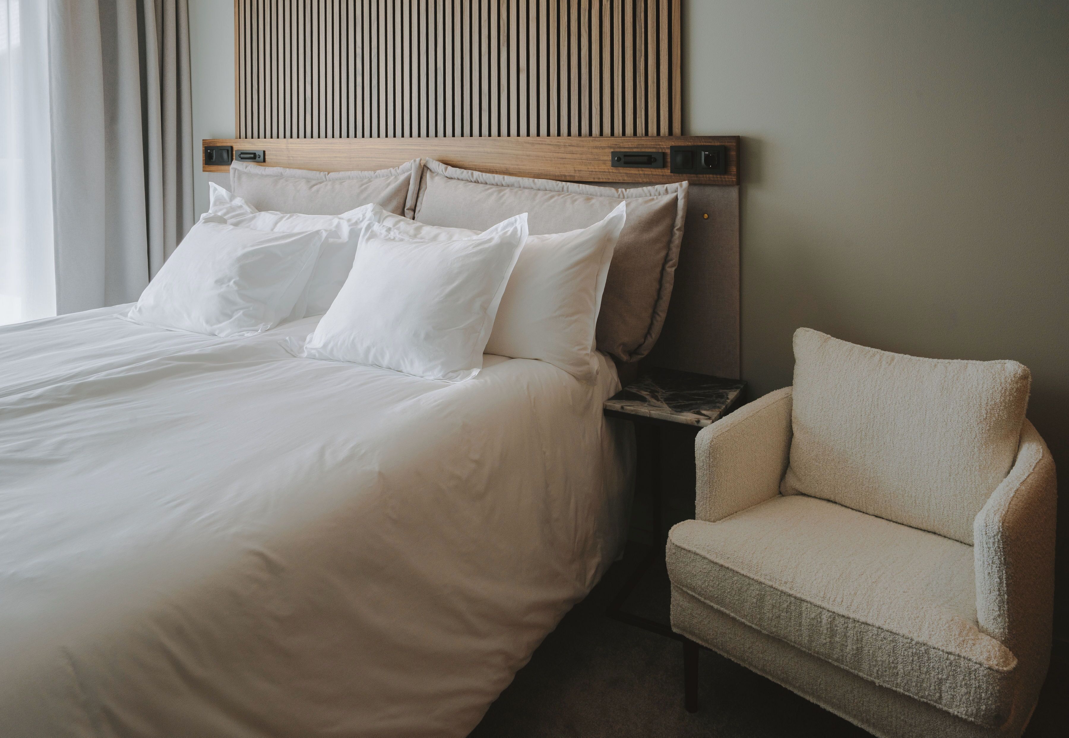 A neatly made bed with white linens and pillows, a wooden headboard, and a beige armchair beside it.