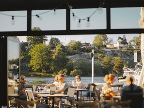 An outdoor dining area with tables and chairs, overlooking a body of water and a rocky hillside with buildings in the background. - Season Hotel (Ekerö)