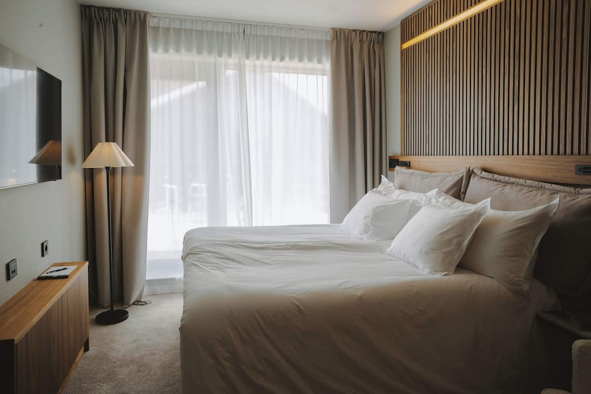 A hotel room with a large bed, a bedside table, a lamp, a TV mounted on the wall, and a window with curtains.