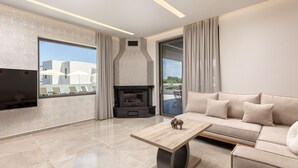 A modern living room with a sofa, a coffee table, a fireplace, and large windows. - Villa Frantzeska (Zakynthos)