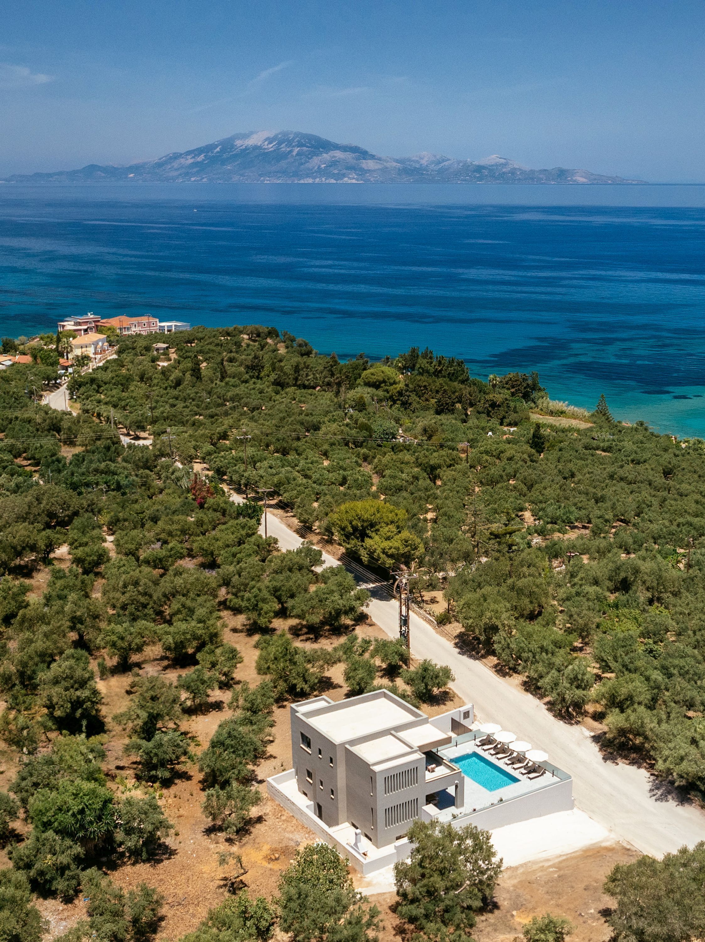 Eine Vogelperspektive zeigt eine moderne Villa mit Schwimmbad, umgeben von üppiger Vegetation und freiem Blick auf das Meer und ferne Berge.