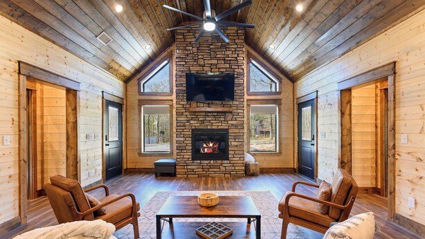 A log cabin interior with a stone fireplace, wooden walls, and a ceiling fan. - The Hickory Nut | 2 Bed | 2.5 Bath | Sleeps 4 (Broken Bow)